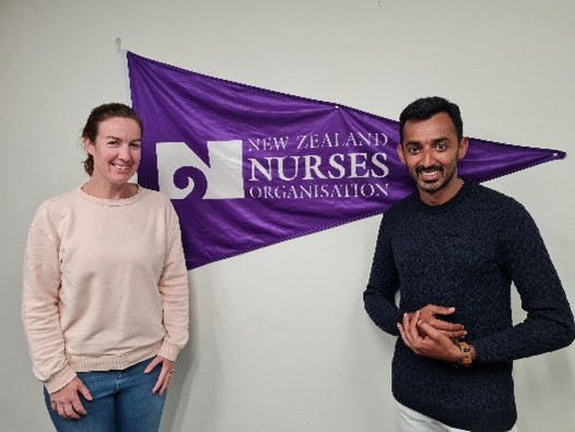 Two smiling people standing in front of a triangular NZNO flag which is mounted n a wall